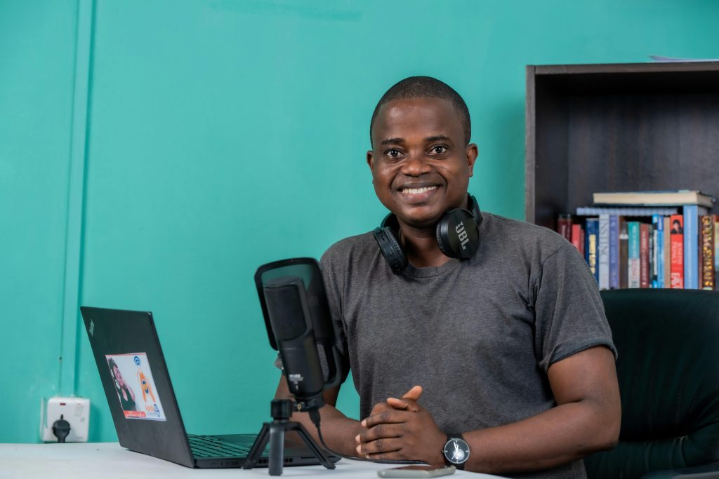 African man smiling in a home studio setting with laptop, microphone, and headphones, ideal for tech or podcast themes.